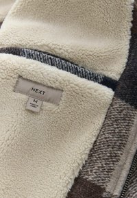 Doublure en polaire douce de couleur crème à l'intérieur d'une veste en laine de taille moyenne avec un motif à carreaux noir, blanc et marron et une étiquette en tissu portant la marque "NEXT."