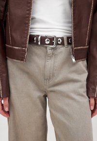 Primer plano de una persona que lleva pantalones beige, camiseta blanca, chaqueta de cuero marrón y cinturón marrón oscuro con hebilla plateada y tachuelas redondas.