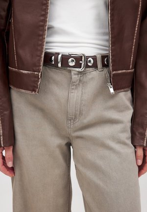 Tuva cilvēka apģērba detaļas: bēša krāsas bikses, balta blūze, brūna ādas jaka un tumši brūns josta ar sudraba sprādzi un apaļiem kniedēm.