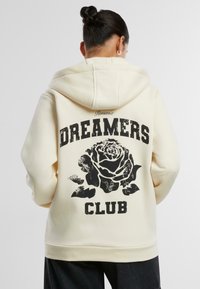 Cremefarbener Hoodie mit einem schwarzen Rosenmotiv und dem Text "DREAMERS CLUB" auf dem Rücken, ausgestattet mit einer Kapuze und gerippten Bündchen.