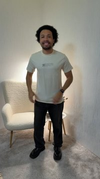 Homme aux cheveux bouclés portant un t-shirt blanc à motif, un jean noir et des chaussures noires, debout en souriant dans une pièce avec un fauteuil blanc et une petite table.