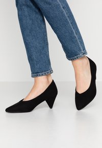 Escarpins à talons hauts en daim noir avec un bout pointu et un talon triangulaire, associés à un jean en denim bleu retroussé.