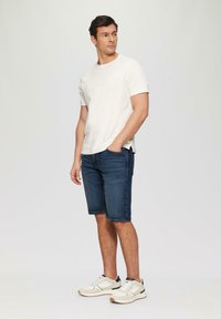 Witte kortemouwen shirt, donkere denim shorts en lichte sportschoenen. De outfit heeft een relaxte pasvorm met minimale naden en geen patronen.