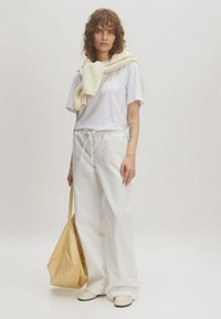 Witte oversized t-shirt, crème trui over de schouders gedrapeerd, witte wijde broek, beige tas en lichtgekleurde instappers.