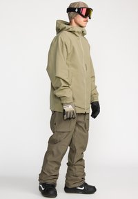Snowboardjas in olijfgroen, met een ritssluiting aan de voorkant, capuchon en een relaxte pasvorm. Gecombineerd met bijpassende cargobroek en zwarte handschoenen.