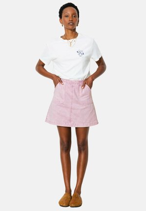 Femme debout, les mains dans les poches, portant un t-shirt blanc, une jupe rose, des pantoufles marron et des bijoux en or, sur un fond uni.