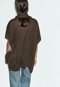 Brun stickad poncho med hög krage, lös passform och sidslitsar, parat med ljusblå jeans. Slät textur, avslappnad design.