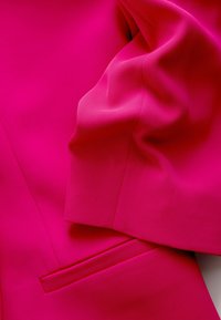 Žiarivo ružová látka s viditeľným švom a detailom vrecka, ktorá ukazuje hladkú a mierne pokrčenú textúru.
