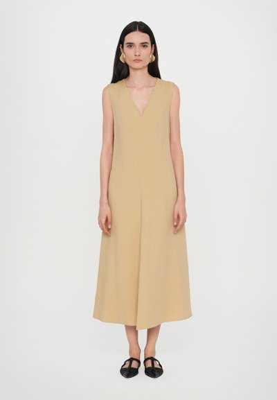 Robe sans manches jaune moutarde avec un col en V, longueur au genou, confectionnée dans un tissu texturé. Portée avec des chaussures plates noires.