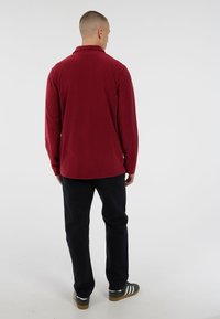 Man staat met zijn rug naar de camera, gekleed in een bordeauxrode langekoope shirt, zwarte broek en grijze Adidas sneakers met witte strepen.