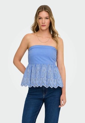 Jeune femme portant un haut bleu bustier avec un ourlet en dentelle ajourée brodée et un jean bleu foncé, debout devant un fond blanc.