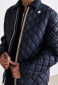 Veste matelassée bleu marine avec motif en losanges, accents blancs, jaunes et rouges le long de la fermeture éclair, et un logo sur la poitrine gauche.