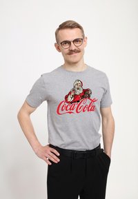 Muž s brýlemi a knírem, oblečený v šedém tričku s postavou Santa Clause, který drží lahvičku Coca-Coly, a s červeným logem Coca-Cola.