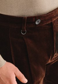 Brązowe spodnie w kordurze o teksturowej powierzchni, z metalowym guzikiem i metalową pętlą w pasie, uzupełnione beżowym swetrem.