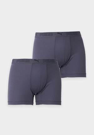 Jeune homme debout pieds nus portant un boxer gris foncé ajusté sur un fond clair uni.