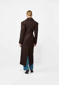 Długi brązowy płaszcz z strukturalnego materiału, z wywiniętym kołnierzem, prostą sylwetką i centralnym rozcięciem z tyłu, zestawiony z niebieskimi dżinsami.