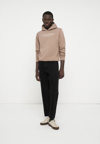 Giovane uomo che indossa un hoodie beige Jack & Jones, jeans neri e sneakers beige, in piedi di fronte a uno sfondo bianco semplice.