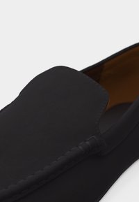 Mocassin noir à enfiler avec bout carré arrondi et surpiqûres visibles le long du bord supérieur, doublure intérieure marron.