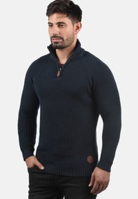 Marineblau gestrickter Pullover mit hohem Kragen und halbem Reißverschluss. Rippstruktur, figurbetontes Design, mit einem braunen Lederlogo-Patch am Saum.