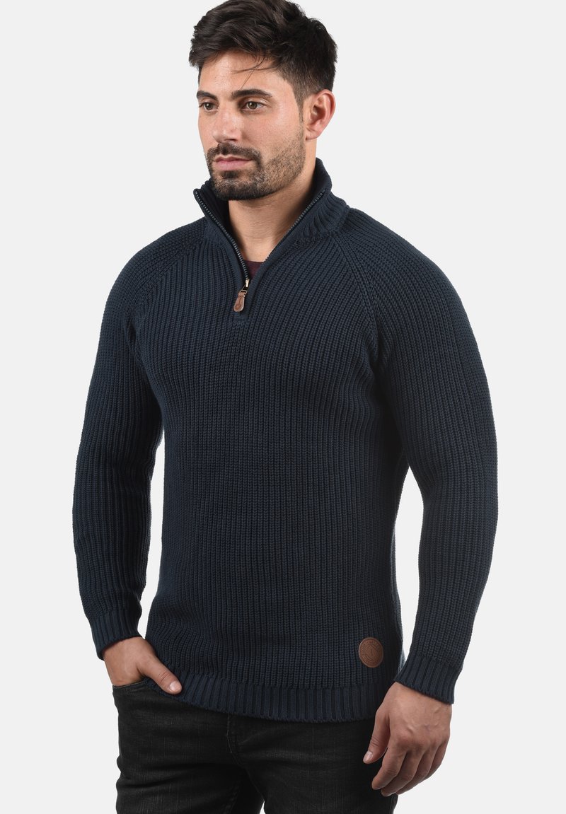 Marineblau gestrickter Pullover mit hohem Kragen und halbem Reißverschluss. Rippstruktur, figurbetontes Design, mit einem braunen Lederlogo-Patch am Saum.