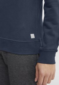 Tamsiai mėlyna džemperis su gipiūro rankogaliais ir apykakle, ant kurio yra mažas baltas etiketė su užrašu "DNM WORKRS." Medžiaga atrodo minkšta ir tekstūruota.