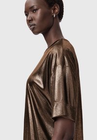 Bronzfärgad metallisk oversized t-shirt med korta ärmar, med en slät, reflekterande yta och en rundad halsringning, visad på en models profil.