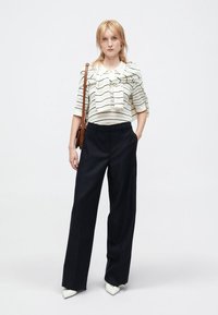 Blusa de rayas blancas y negras con escote de lazo, combinada con pantalones de talle alto en azul marino y zapatos de punta blanca. Bolso de hombro marrón.