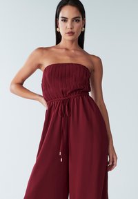 Vijoličen kombinezon brez naramnic z navpičnimi gubami, elastičnim pasom z zateznim trakom in širokimi hlačnicami, narejen iz gladkega blaga.