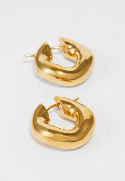 Paire de grosses boucles d’oreilles créoles dorées brillantes aux formes irrégulières légèrement torsadées sur fond blanc.
