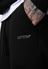 Schwarze Jogginghosen mit sichtbar aufgedrucktem Logo "ESTEEM" in Pink und Weiß. Weicher Stoff, elastischer Bund und seitliche Taschen. Texturierte Haut sichtbar.
