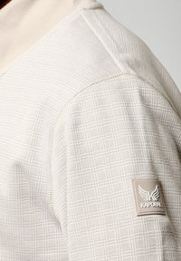 Suéter de color beige claro con un patrón texturizado, que cuenta con un cuello alto y un parche con logo en el hombro izquierdo. Acabado de tejido suave.