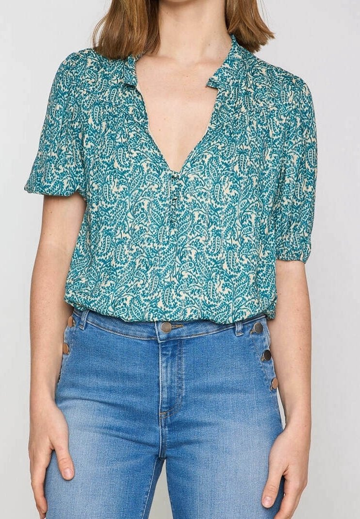 Blusa teal a maniche corte con motivo floreale, scollatura a V e dettagli arruffati sulle spalle, abbinata a jeans blu a vita alta.