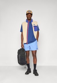 Man in een beige fleece vest, blauwe shirt, lichtblauwe shorts en een tan pet, die een zwarte rugzak vasthoudt tegen een effen grijze achtergrond.
