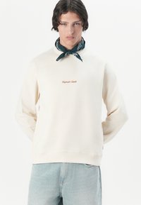En model bærer en cremet sweatshirt med et lille logo, parret med et mørkegrønt bandana om halsen, sat mod en hvid baggrund.