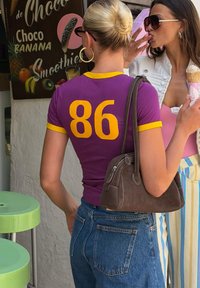 Deux femmes près d'un panneau de smoothie ; l'une porte un t-shirt violet avec le numéro 86, l'autre porte des lunettes de soleil et mange un cornet de glace.