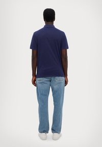 Námořnická modrá polo tričko s krátkými rukávy a límcem, v kombinaci s světle modrými džíny a bílými teniskami. Pohled zezadu.