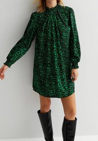 Zelené šaty s leopardím potiskem, dlouhými nabíranými rukávy a nabíraným výstřihem, spárované s černými kozačkami do kolen. Hladká textura, volný střih.