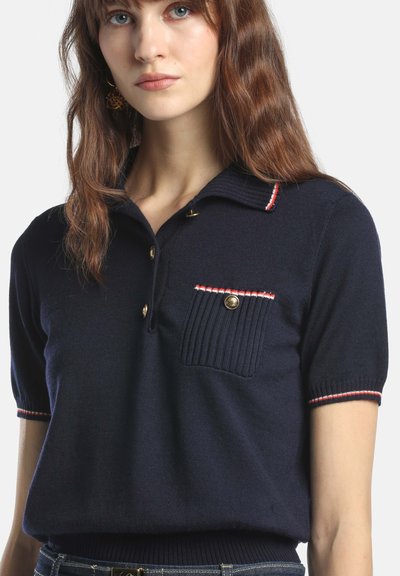 Tricou polo bleumarin cu mâneci scurte, guler striat, trei nasturi de aur și o buzunare pe piept accentuată cu bordură roșie și albă.