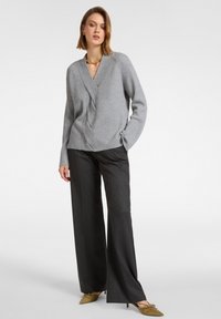 Szary sweter prążkowany z dekoltem w serek i asymetrycznym wzorem, zestawiony z czarnymi spodniami o szerokich nogawkach i oliwkowozielonymi szpilkami.