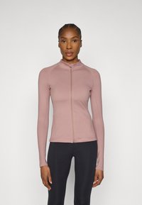 Long-sleeve, zip-up top in light pink, made of smooth fabric with a high collar and flat seams. Paired with black leggings.