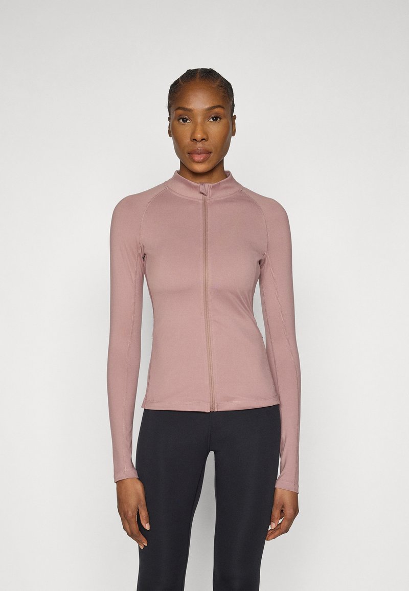 Long-sleeve, zip-up top in light pink, made of smooth fabric with a high collar and flat seams. Paired with black leggings.