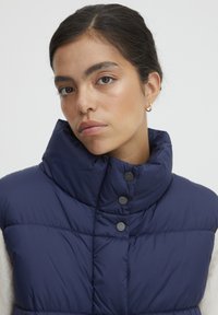 Vest puffer de marină cu guler înalt, dotat cu butoane tip capse și textură matlasată. Proiectat pentru căldură, având o siluetă elegantă și aerodinamică.