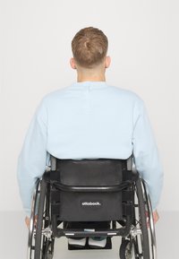 Personne aux cheveux châtain clair portant un sweat-shirt bleu clair assise dans un fauteuil roulant noir Ottobock, vue de dos.