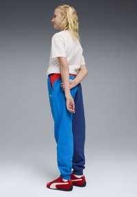Blaue und marineblaue Jogginghosen mit einem roten Bund, kombiniert mit einem weißen T-Shirt und roten Turnschuhen. Die Textur wirkt weich; sichtbare Logodetails.