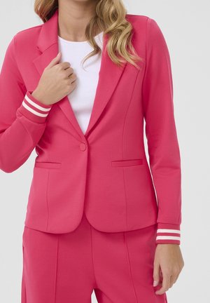 Femme portant une veste et un pantalon de costume rose vif avec des poignets rayés blancs, sur une chemise blanche unie, main ajustant le revers de la veste.