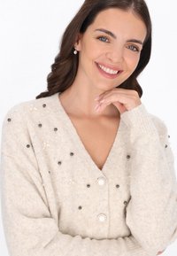 Beżowy sweter z dzianiny, z dekoltem w kształcie V, ozdobiony perłami i koralikami. Posiada dwa zapięcia na guziki i ściągacze przy mankietach. Miękka faktura.