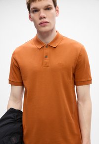Jauns vīrietis, tērpts apdedzināti oranžā polo krekliņā ar "BOSS" logotipu uz krūtīm, kreisajā rokā tur melnu jaku uz viendabīga fona.
