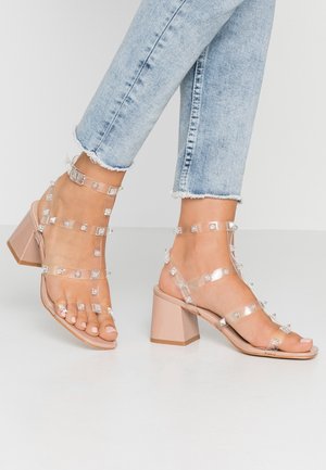 Przezroczyste sandały na obcasie z przezroczystymi paskami ozdobionymi kwadratowymi ćwiekami, z cielistym słupkowym obcasem i otwartym noskiem.