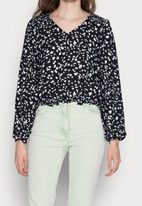 Schwarze Bluse mit langen Ärmeln und weißem Sprenkelmuster, V-Ausschnitt, elastischem Bund, kombiniert mit mintgrünen Cordhosen.
