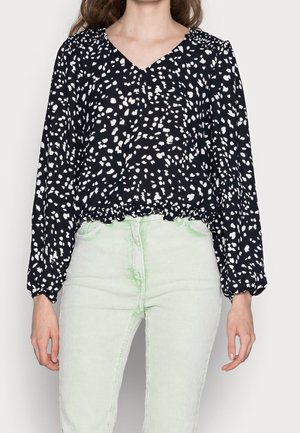 Schwarze Bluse mit langen Ärmeln und weißem Sprenkelmuster, V-Ausschnitt, elastischem Bund, kombiniert mit mintgrünen Cordhosen.